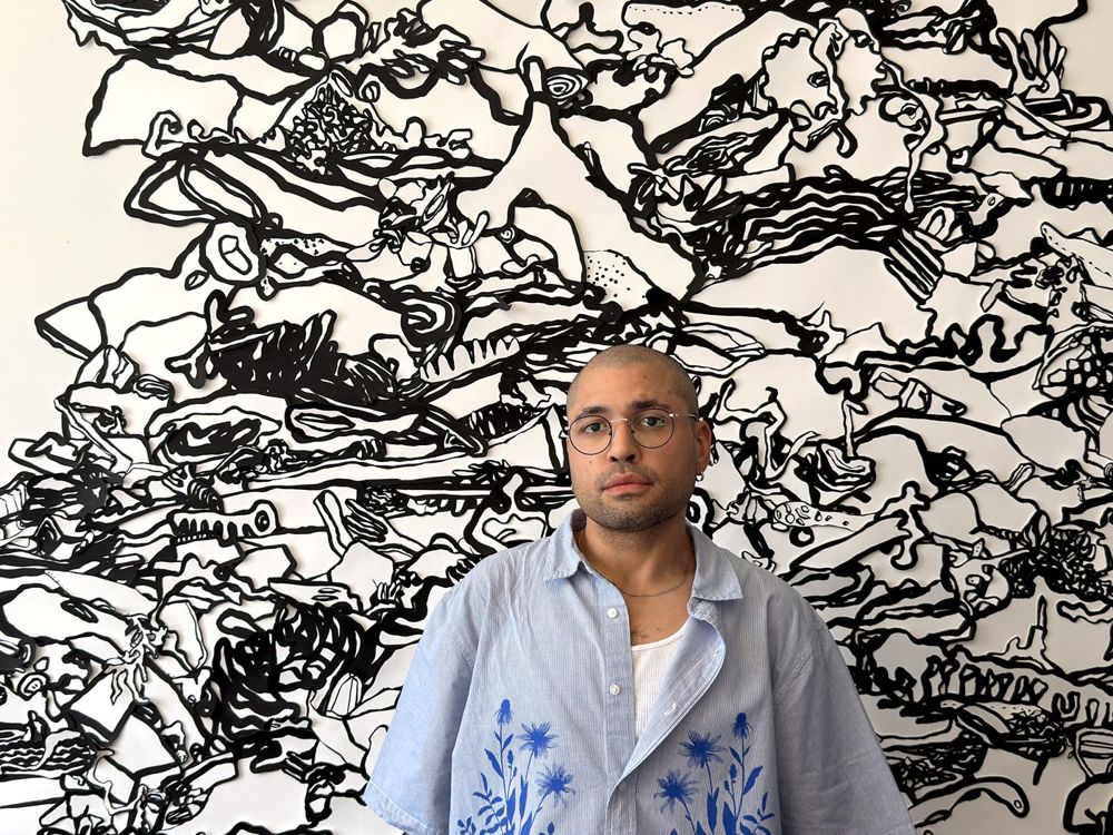 Zach Blackwood standing in front of a black and white abstract mural, wearing glasses and a light blue button-up shirt with blue floral print.